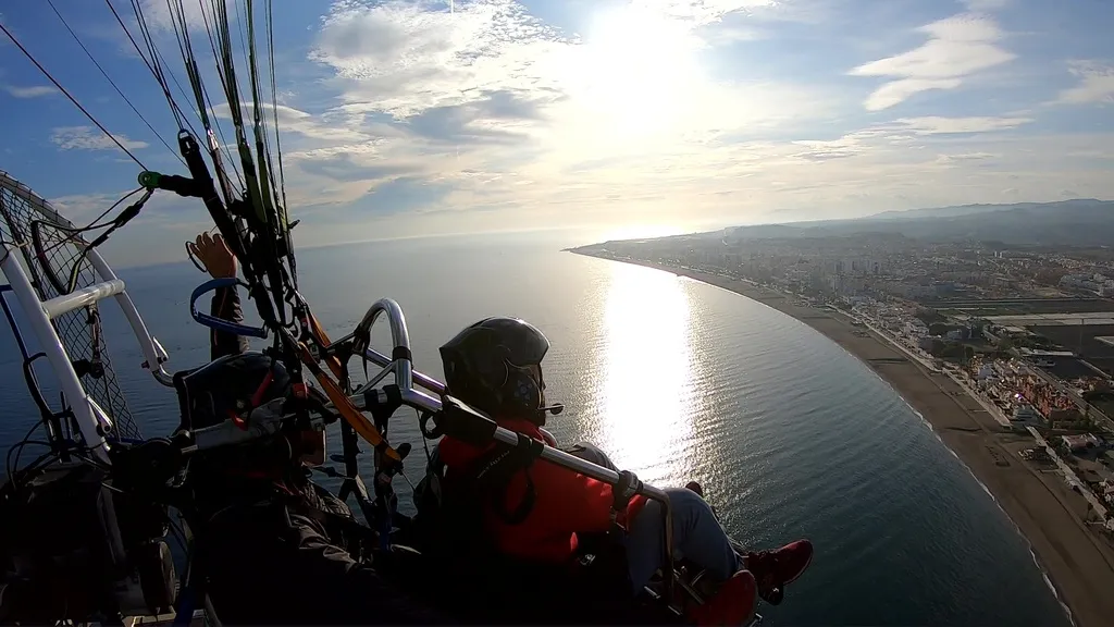 Dos personas en un vuelo en paramotor biplaza, sobrevolando la costa con el reflejo del sol sobre el mar.