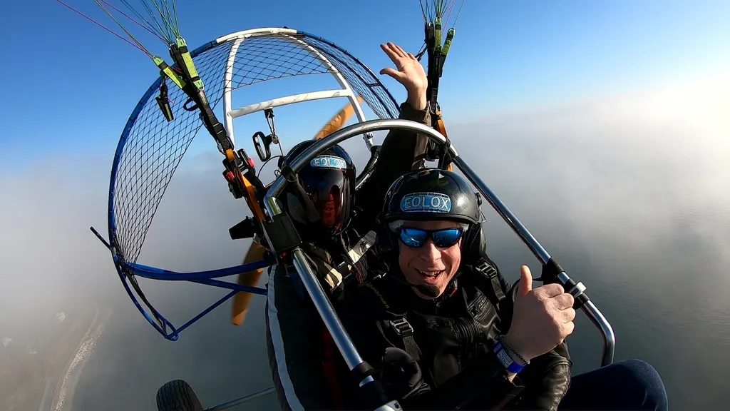 Pasajero sonriente con pulgar arriba en un selfie durante un vuelo en paramotor biplaza sobre un mar de nubes.