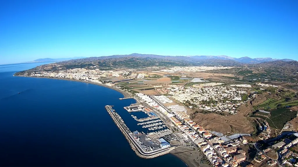 Vista aérea desde un vuelo en paramotor de una ciudad costera con un puerto deportivo y montañas al fondo bajo un cielo azul.