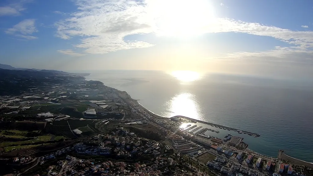 Vista panorámica desde un vuelo en paramotor de la costa, un puerto deportivo y campos, con el sol brillando sobre el mar.