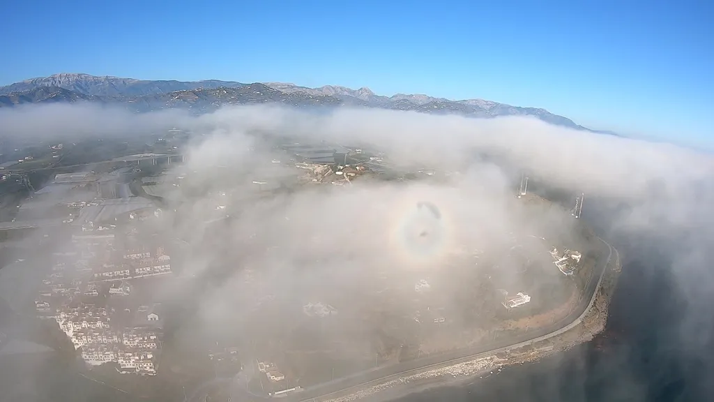 Vuelo en paramotor a través de las nubes, mostrando un halo de luz o espectro de Brocken sobre un paisaje cubierto de niebla.